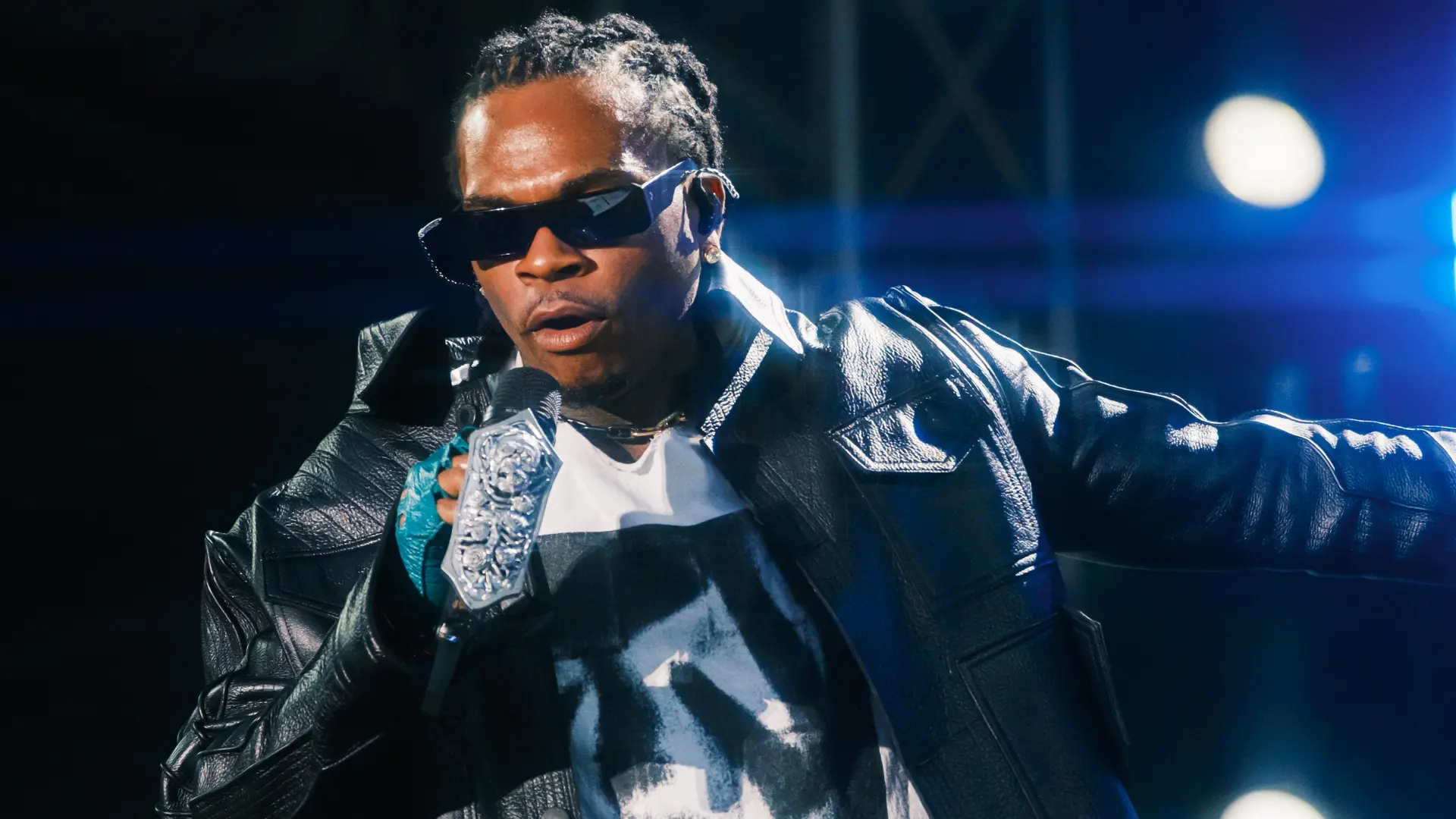 Rapper Gunna performing on stage, wearing sunglasses and a sleeveless shirt, smiling while holding a mic.