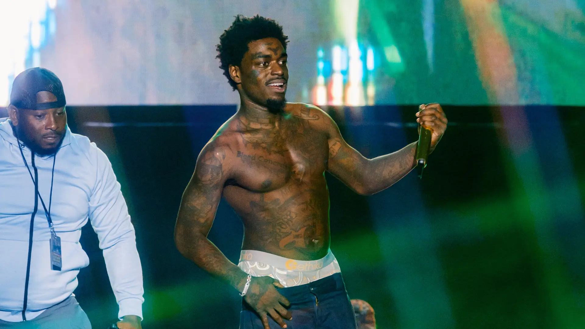 Rapper Kodak Black performs shirtless on stage, holding a microphone with a crew member nearby.